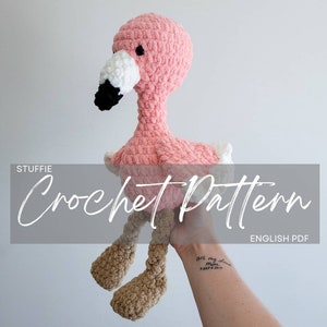 May include: Pink and brown crocheted flamingo stuffed animal. The crochet pattern is available as an English PDF download.  The stuffed animal is being held by a hand.