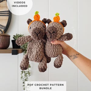 Peut inclure: Deux peluches capybara au crochet marron en fil texturé, portant chacune un petit chapeau citrouille orange avec une tige verte. L'image contient le texte HELPFUL VIDEOS INCLUDED et PDF CROCHET PATTERN BUNDLE. Les jouets ont des yeux et nez foncés.