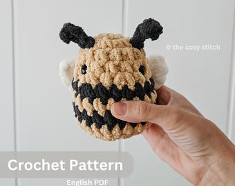 Pode incluir: Padrão de crochê para um brinquedo amigurumi de abelha. A abelha é marrom e preta com asas brancas. O padrão está disponível em formato PDF em inglês.