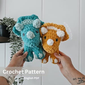 May include: Two crocheted triceratops dinosaurs, one teal and one mustard yellow, are held up. Each has white accents and black eyes. The text "Crochet Pattern" and "English PDF" are at the bottom of the image.