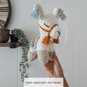 May include: A white crocheted llama plush toy with light blue tassels on its ears, a mustard yellow bridle, and a matching saddle. The llama has closed eyes and light brown feet. The text PDF CROCHET PATTERN is visible at the bottom of the image.