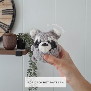 Pode incluir: Um brinquedo de guaxinim de crochê cinza e branco com detalhes em preto. O texto "PDF CROCHET PATTERN" é exibido em um retângulo branco. O brinquedo é segurado na mão.