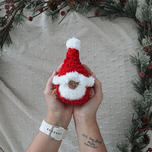 Pode incluir: Uma boneca de gnomo de croch&ecirc; feita &agrave; m&atilde;o em vermelho, branco e marrom. O gnomo tem um chap&eacute;u vermelho com um pompom branco, uma barba branca e um rosto marrom. A boneca &eacute; segurada em duas m&atilde;os, com um rel&oacute;gio branco no pulso. Decora&ccedil;&otilde;es de Natal.