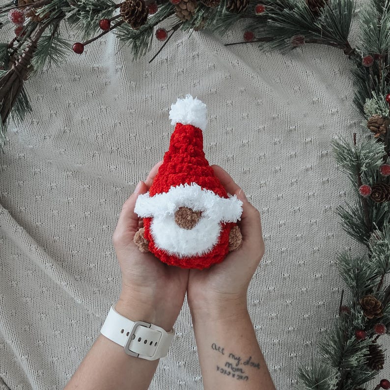 Pode incluir: Uma boneca de gnomo de croch&ecirc; feita &agrave; m&atilde;o em vermelho, branco e marrom. O gnomo tem um chap&eacute;u vermelho com um pompom branco, uma barba branca e um rosto marrom. A boneca &eacute; segurada em duas m&atilde;os, com um rel&oacute;gio branco no pulso. Decora&ccedil;&otilde;es de Natal.