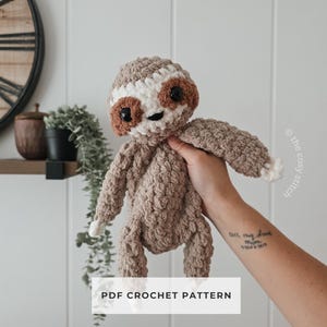 May include: A crocheted sloth toy in brown, white, and beige. The sloth features a textured body, small black eyes, and a smiling expression. The image includes the text "PDF CROCHET PATTERN".