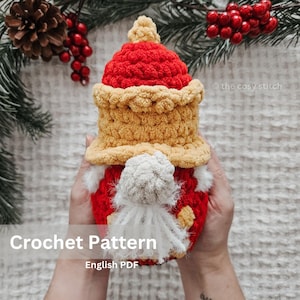 Może przedstawiać: Ręcznie robiony szydełkowy gnom z czerwonym kapeluszem i korpusem, żółtym rondem i białą brodą. Gnom trzyma Crochet Pattern English PDF. The cozy stitch znajduje się w prawym górnym rogu. Świąteczna dekoracja.