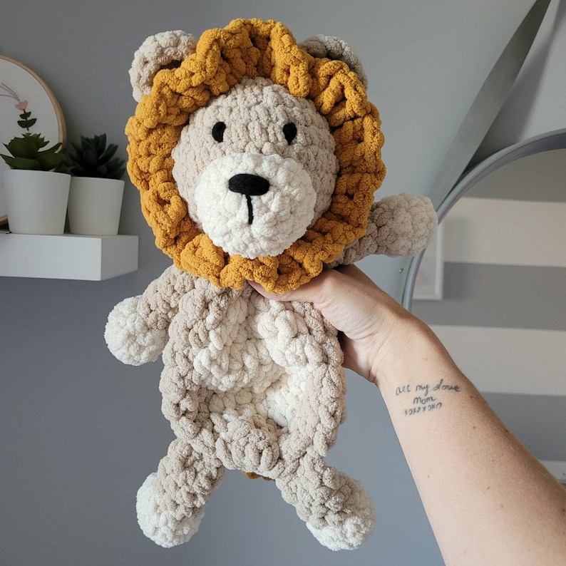 May include: A crocheted lion stuffed animal with a beige body and a yellow mane. The lion is being held by a hand with a tattoo that says "all my love mom" and a date.