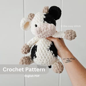 May include: Crochet pattern for a black and white cow amigurumi toy. The pattern is available as an English PDF download. The toy is made with white and black yarn and has a pink nose.