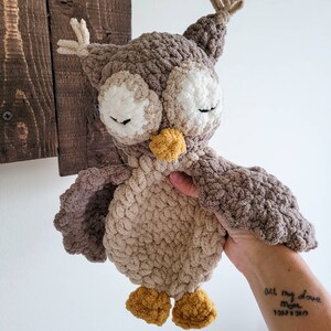 May include: A crocheted owl plush toy with brown and beige fur, closed eyes, and a yellow beak. The owl is being held by a hand with a tattoo that says "All my love Mom" and a date.