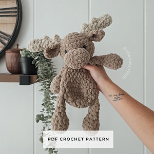 May include: A handmade, plush moose toy in shades of brown and tan. The stuffed animal has large antlers, a round body, and long legs. The text "PDF CROCHET PATTERN" is displayed on a white rectangle below the moose.