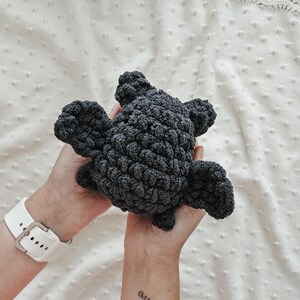 May include: A dark gray crocheted plush toy shaped like a bat with large, rounded ears. The toy is held in the hands of a person.