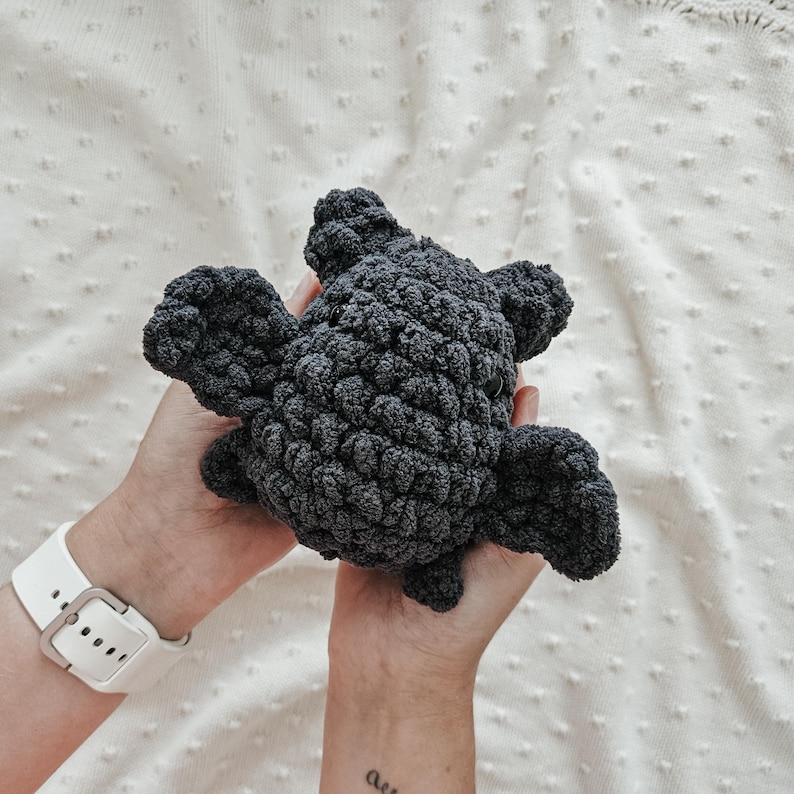 May include: A dark gray crocheted plush toy shaped like a bat with large, rounded ears. The toy is held in the hands of a person.