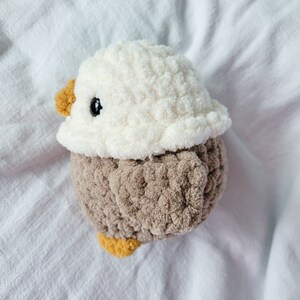 Puede incluir: Un juguete de p&aacute;jaro de peluche tejido a crochet con cabeza blanca y cuerpo marr&oacute;n. El p&aacute;jaro tiene un ojo negro y un peque&ntilde;o pico amarillo.
