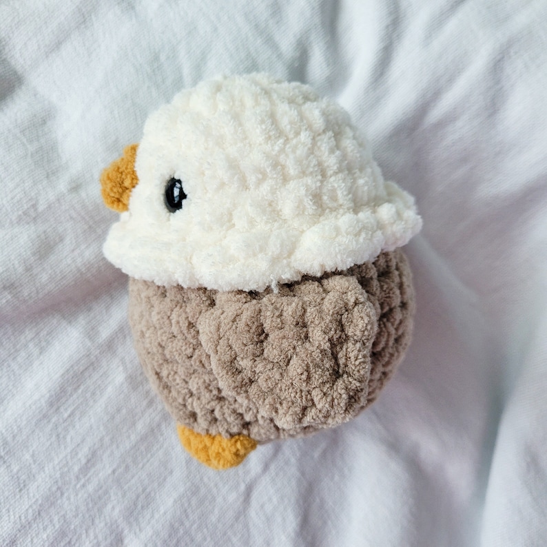 Puede incluir: Un juguete de p&aacute;jaro de peluche tejido a crochet con cabeza blanca y cuerpo marr&oacute;n. El p&aacute;jaro tiene un ojo negro y un peque&ntilde;o pico amarillo.