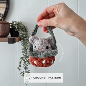 May include: A handmade crochet basket in shades of gray, green, and rust, holding a small gray bunny. The basket has white polka dots and a green trim. The text "PDF CROCHET PATTERN" is displayed.