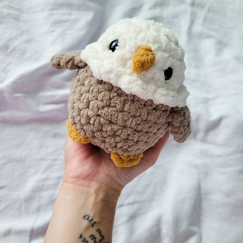Puede incluir: Un juguete de peluche de &aacute;guila tejido a crochet con pelaje marr&oacute;n y blanco. El &aacute;guila tiene una cabeza blanca con ojos negros y un pico amarillo.