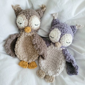 May include: Two crocheted owl toys, one brown and one purple, with closed eyes and a fluffy texture. The owls are lying on a white bedspread.