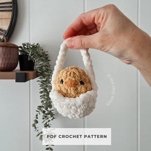 May include: A crocheted chick in a white basket. The chick is light brown with an orange beak and black eyes. The basket has a fluffy white texture and a white handle. The text "PDF CROCHET PATTERN" is at the bottom.