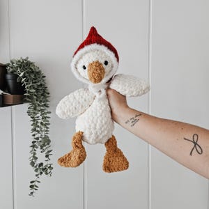 May include: White crocheted goose plush toy wearing a red crocheted Santa hat. The goose has brown felt feet and a brown felt beak.