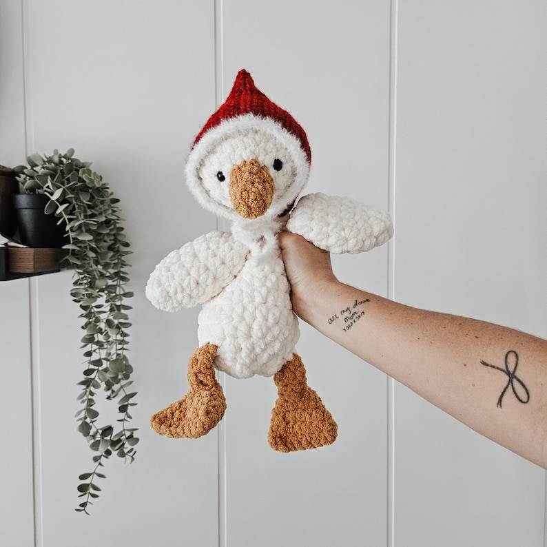 May include: White crocheted goose plush toy wearing a red crocheted Santa hat. The goose has brown felt feet and a brown felt beak.