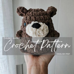 May include: A crocheted brown bear plush toy with white and black facial features. The toy is being held by a hand. The text "Chubby Buddy Crochet Pattern ENGLISH PDF" is displayed on a gray background.