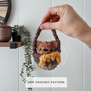 May include: A crocheted bear in a basket with a yellow scarf. The bear is brown and the basket is a darker brown. The text "PDF CROCHET PATTERN" is displayed on a white rectangle at the bottom of the image.