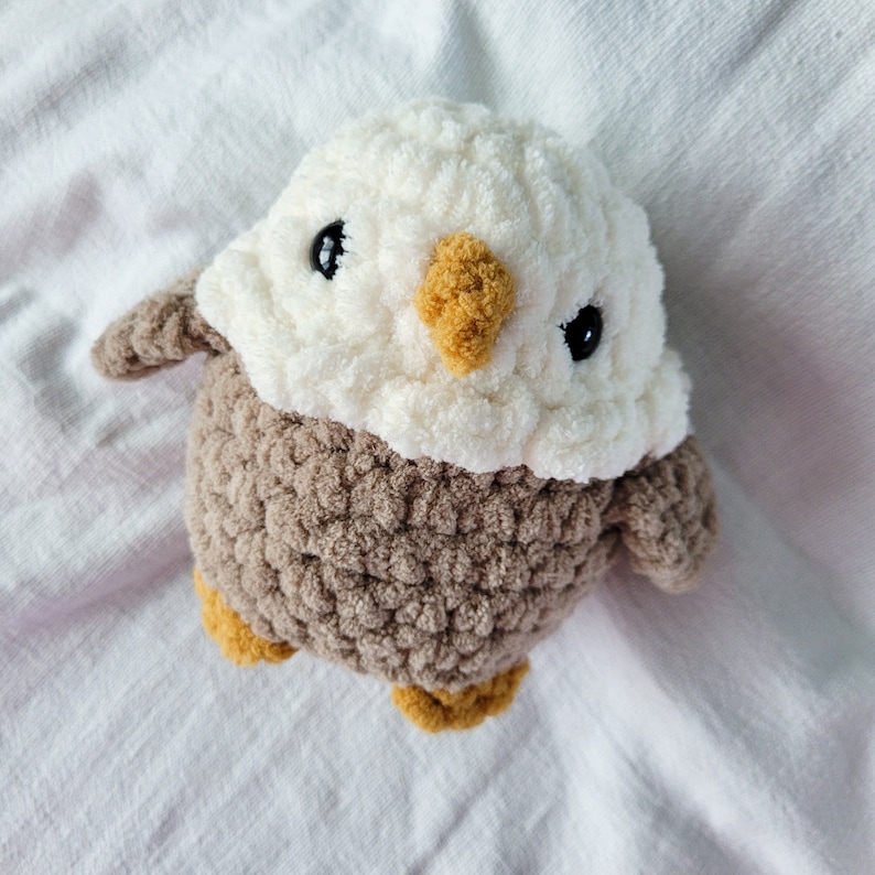 Puede incluir: Un animal de peluche de crochet con forma de &aacute;guila hecho con hilo marr&oacute;n y blanco. El &aacute;guila tiene ojos negros y un pico amarillo.