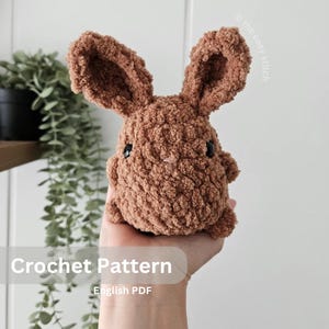 May include: A crochet pattern for a brown bunny amigurumi toy. The pattern is available as an English PDF download. The image shows a completed bunny toy.