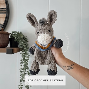 May include: A handmade crochet donkey toy in shades of gray, white, and black. The donkey has large ears, a fluffy mane, and a colorful scarf. The text "PDF CROCHET PATTERN" is displayed below the donkey.
