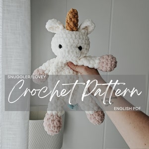Puede incluir: Un juguete de peluche de unicornio de crochet con cuerpo blanco, pezuñas rosas y un cuerno marrón. El juguete está siendo sostenido por una mano y el texto "Snuggler/Lovey Crochet Pattern English PDF" es visible en la imagen.