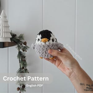 May include: A crocheted penguin with a black head, white face, grey body, and yellow beak and feet. The image includes the text "Crochet Pattern" and "English PDF".