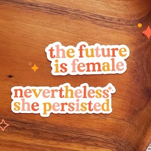 Puede incluir: Dos pegatinas con lemas feministas sobre una superficie de madera. La pegatina superior dice "the future is female" en letras naranja, rosa y amarillo. La pegatina inferior dice "nevertheless she persisted" en letras naranja, rosa y amarillo.