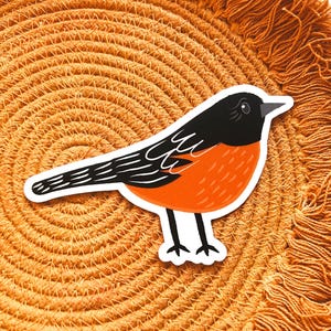 May include: A sticker featuring a stylized bird with a black head and wings, and an orange body. The bird is perched on a textured, orange, circular surface. The sticker has a white border.