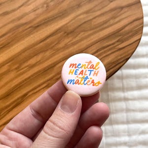 May include: A round, light pink button with the words "mental HEALTH matters" in colorful, handwritten-style lettering. The button is held in a hand, with a wooden surface in the background.