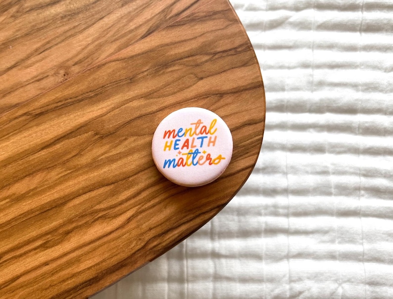 May include: A round, light pink button with the words "mental health matters" in colorful, handwritten-style lettering. The button is resting on a wooden surface with a natural wood grain pattern.