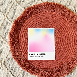 Square sticker with a gradient of pink, blue, and yellow above the words Cruel Summer and the hex codes FDADD6, 68B4DA, and FDECB7. The sticker is on a round, pink woven mat