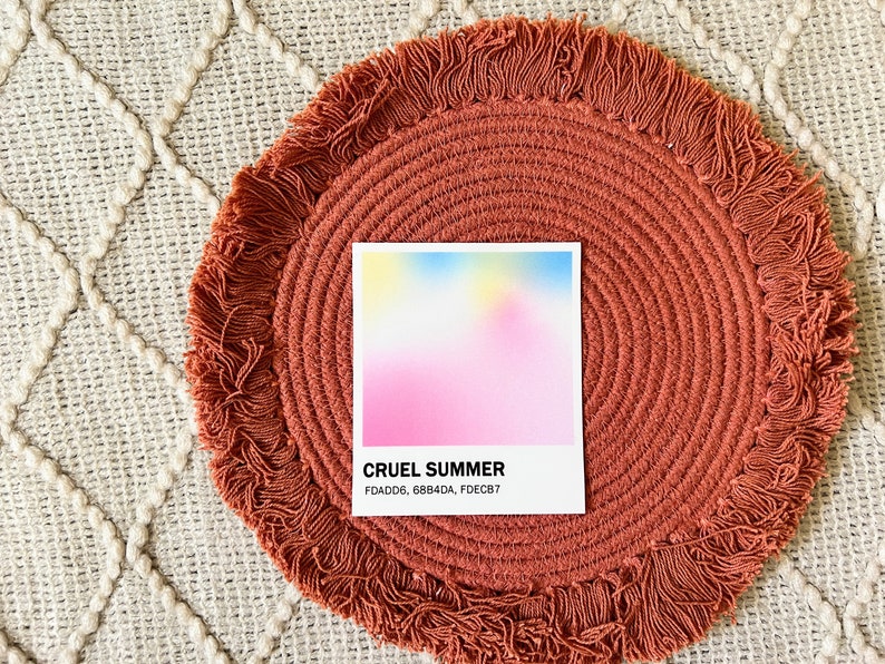 Square sticker with a gradient of pink, blue, and yellow above the words Cruel Summer and the hex codes FDADD6, 68B4DA, and FDECB7. The sticker is on a round, pink woven mat
