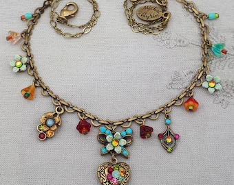 Può includere: Una collana a catena color bronzo con chiusura a moschettone e un'etichetta rotonda con la scritta Michal Negrin. La collana è decorata con vari ciondoli colorati, tra cui piccoli fiori, una farfalla e un pendente a forma di cuore.