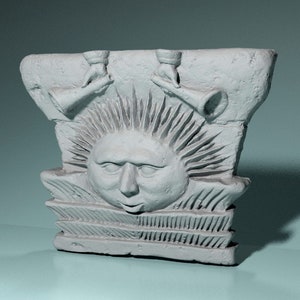 Pode incluir: Uma escultura em relevo de cerâmica branca de um rosto de sol com um halo espinhoso. Duas figuras com chifres estão tocando trombetas acima do rosto do sol.