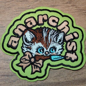 May include: Embroidered patch featuring a cartoon kitten with blue eyes, brown and white fur, and a bow. The patch has the text "march is" in a green, wavy border.