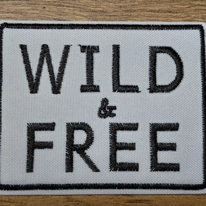 May include: A rectangular patch with the words "WILD & FREE" embroidered in black on a white background. The patch has a black border and is likely designed to be sewn onto clothing or accessories.