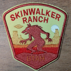 May include: Embroidered patch featuring a red Skinwalker creature with a desert landscape, a UFO, and the text "SKINWALKER RANCH UTAH" in red and gold. The patch has a light yellow background and a red border.