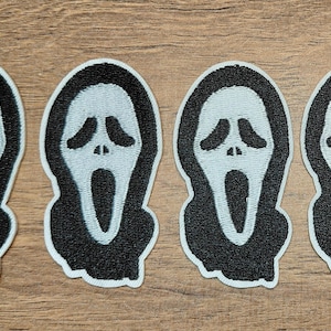 May include: Four embroidered patches featuring the iconic Scream mask design. Each patch has a white face with black eyes and mouth, outlined in black. The patches are on a wooden surface.