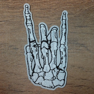 May include: Embroidered patch featuring a white skeleton hand in a rock and roll gesture. The patch has a black outline and is set against a wooden background. The design is detailed and suitable for jackets, bags, or other fabric items.