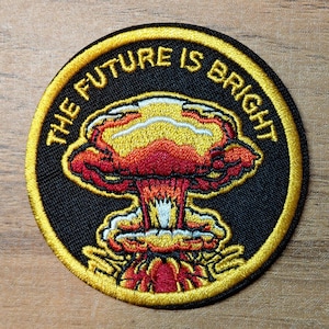 May include: Round embroidered patch with the text "THE FUTURE IS BRIGHT" in yellow. The patch features a colorful mushroom cloud design in red, yellow, and white against a black background, with a yellow border.