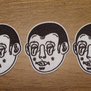 May include: Three embroidered patches featuring a stylized face with large, oval eyes, a small mouth, and dark hair. The patches are primarily white with black detailing and are arranged horizontally.
