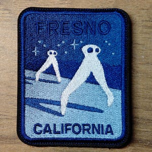 May include: Embroidered patch featuring a blue and white design with the text "FRESNO CALIFORNIA." The patch depicts two white figures with star accents against a gradient blue background. The patch has a dark blue border.