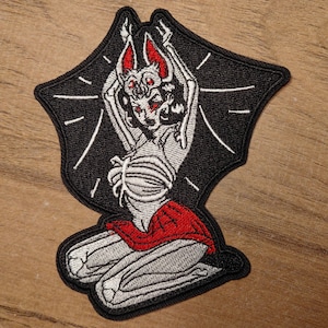 May include: Embroidered patch featuring a skeletal bat-like figure with red accents, kneeling with arms raised. The figure is set against a black bat-wing shaped background with white radiating lines. The patch has a red skirt.