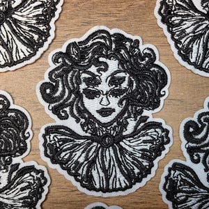 May include: Embroidered patches featuring a stylized woman's face with multiple eyes and elaborate hair. The patches are black and white, with a detailed design and a scalloped edge. The patches are likely for clothing or accessories.