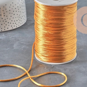 Può includere: Una bobina di cordino in raso giallo oro vibrante avvolto strettamente attorno a un cilindro di plastica bianca. Il cordino liscio e lucido si estende dalla bobina, formando un cappio sciolto. Il materiale ha una consistenza setosa e riflettente.